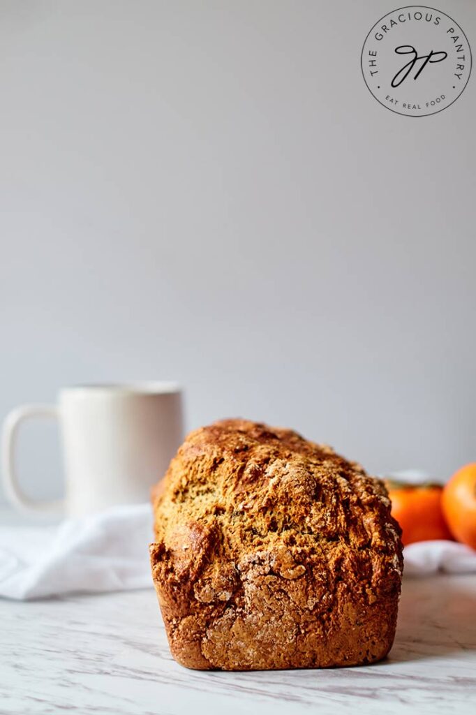 A whole loaf of persimmon bread sits, ready to be sliced on a marble counter top.