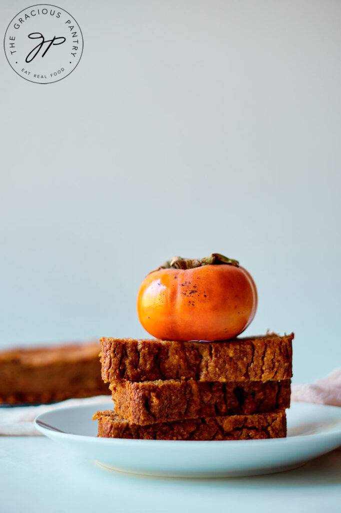 Three thick slices of persimmon bread are stacked on a plate with a persimmon sitting on top.