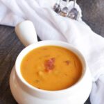 A bowl of this Bacon Sweet Potato Soup sits on a table with a white towel to the side and some measuring spoons in the background.