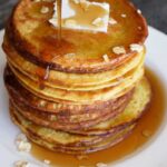 A stack of oat pancakes with maple syrup being poured over the top.