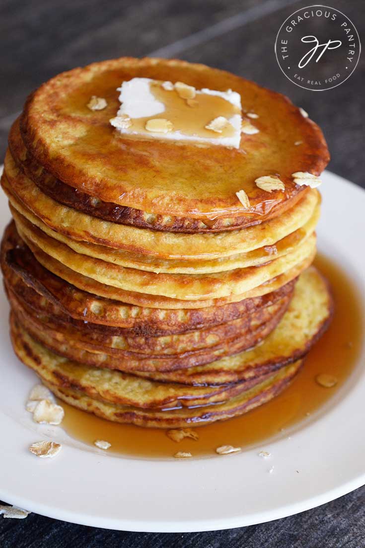 A stack of warm, oat pancakes on a white plate on a dark background. Butter and maple syrup top the pancakes.