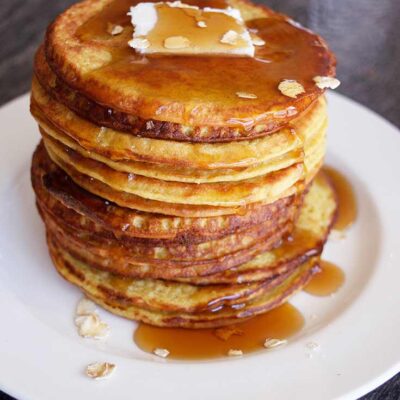 A stack of oat pancakes on a white plate, topped with a pat of butter and plenty of maple syrup.