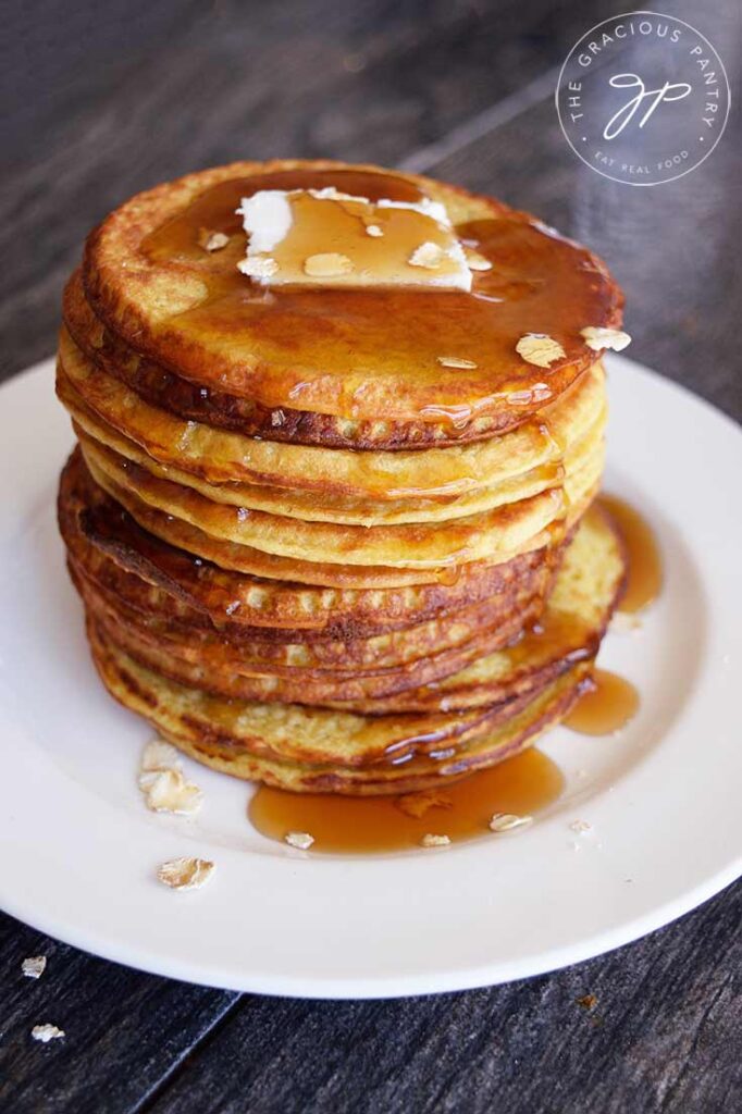 A stack of oat pancakes on a white plate, topped with a pat of butter and plenty of maple syrup.