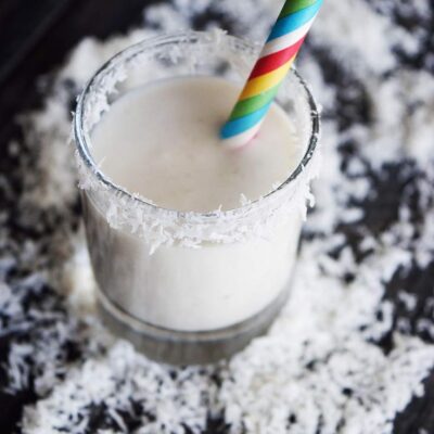 A shot glass filled with this Coconut Milk Smoothie, surrounded by coconut shreds.