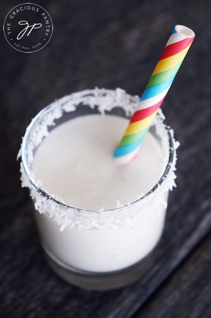 A coconut milk smoothie in a shot glass with a rainbow straw and coconut shreds around the rim of the glass.