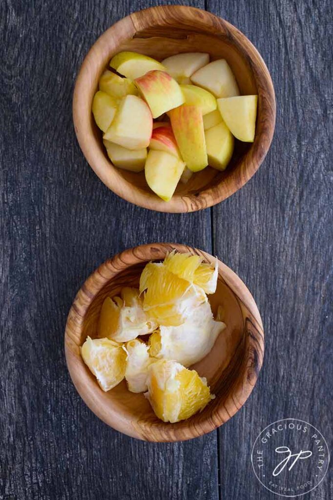 Two wooden bowls, one filled with chopped apples, the other filled with chopped oranges, for making cranberry relish.