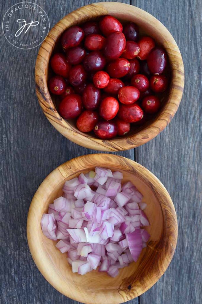 Two wooden bowls, one filled with fresh cranberries, and the other with chopped red onions. Both ingredients are used in making this cranberry relish.