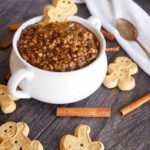 A bowl of gingerbread oatmeal sits on a table surrounded by gingerbread man cookies and cinnamon sticks.