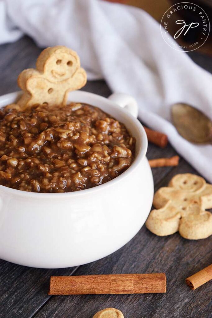 A side view of a white bowl filled with gingerbread oatmeal. Cinnamon sticks and cookies are scattered around the bowl.