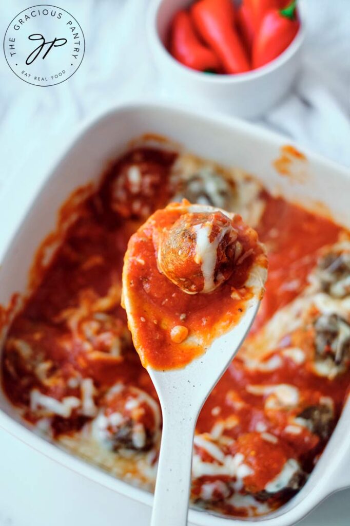One of the cheesy baked meatballs on a serving spoon held over the casserole dish it's being served from.