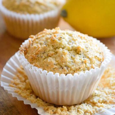 A Gluten Free Lemon Poppy Seed Muffin sits on an unfolded cupcake paper with a lemon behind it.