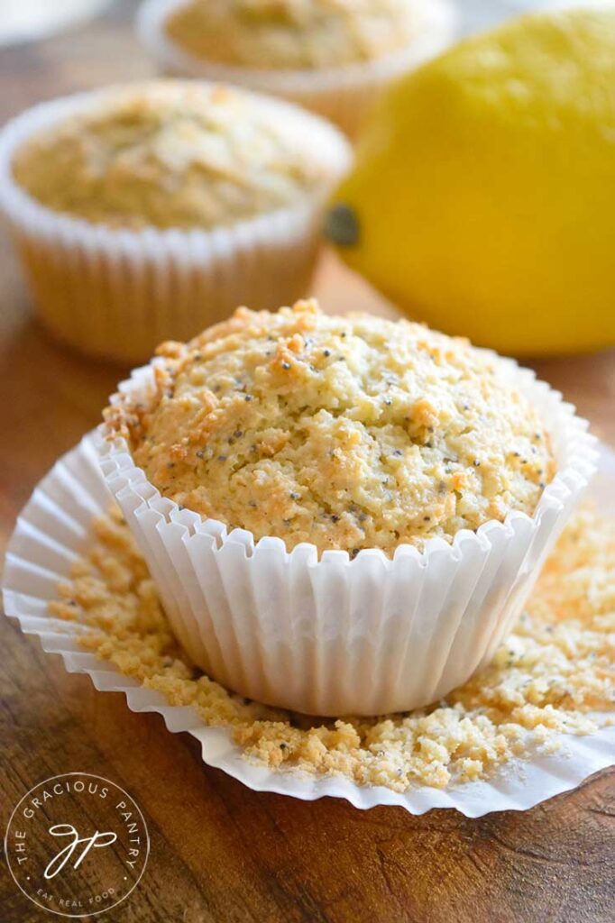 A Gluten Free Lemon Poppy Seed Muffin sits on an unfolded cupcake paper with a lemon behind it.