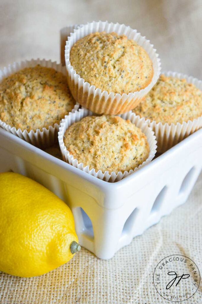 Gluten Free Lemon Poppy Seed Muffins stacked in a white, square, ceramic basket with a lemon sitting beside it.