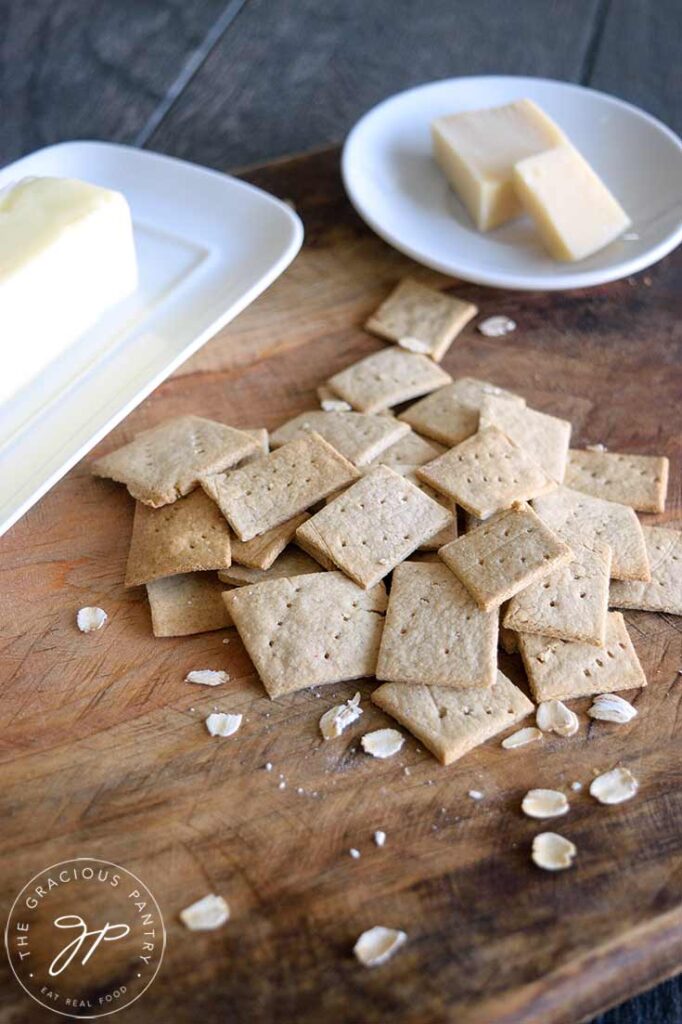 Oat crackers on a cutting boar with a butter dish and a cheese dish sitting behind them.