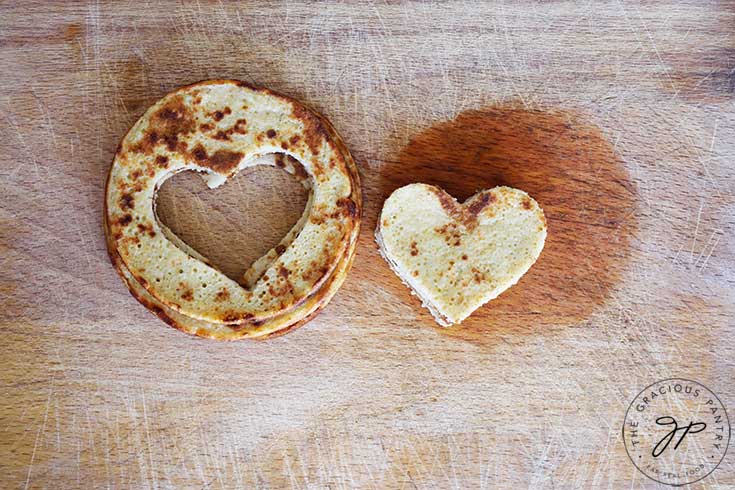 Two piles of pancakes sit next to each other on a cutting board. On the left are the regular pancakes with hearts cut out of the center. On the right are the cut out hearts for a Valentine Pancake Sandwich.