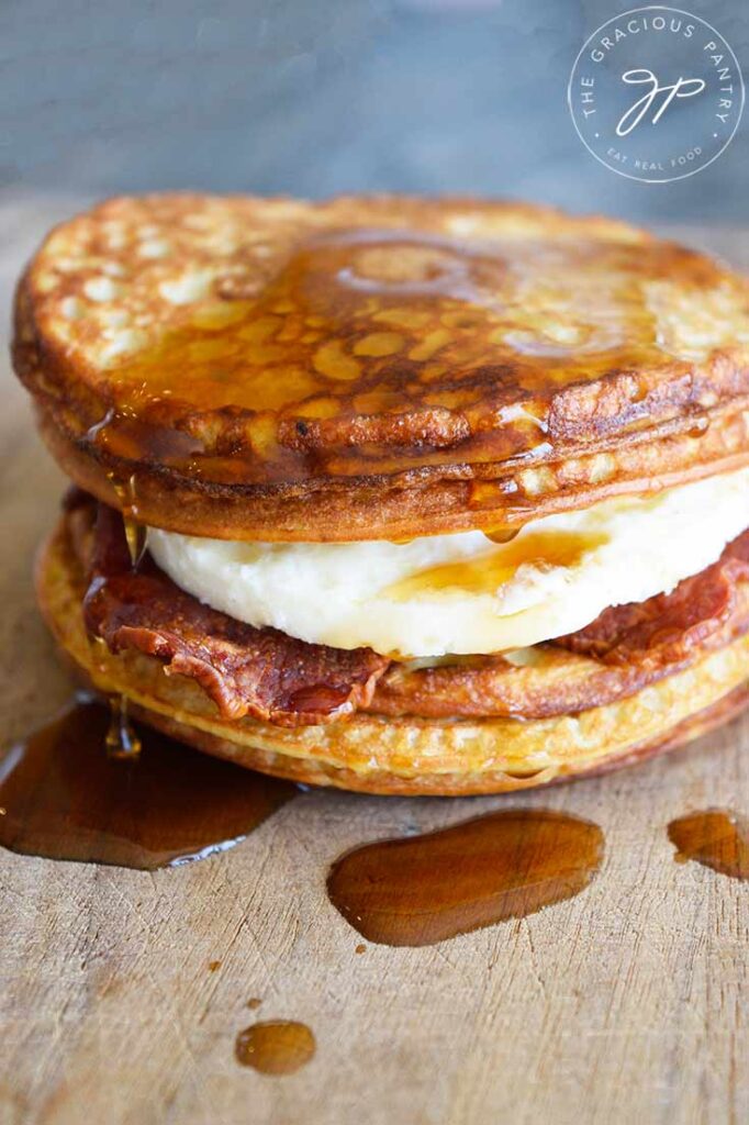 A stack of four pancakes sit on a cutting board. Between them are pieces of bacon and a fried egg. They are put together like a sandwich and have maple syrup dripping over the top, down onto the cutting board.