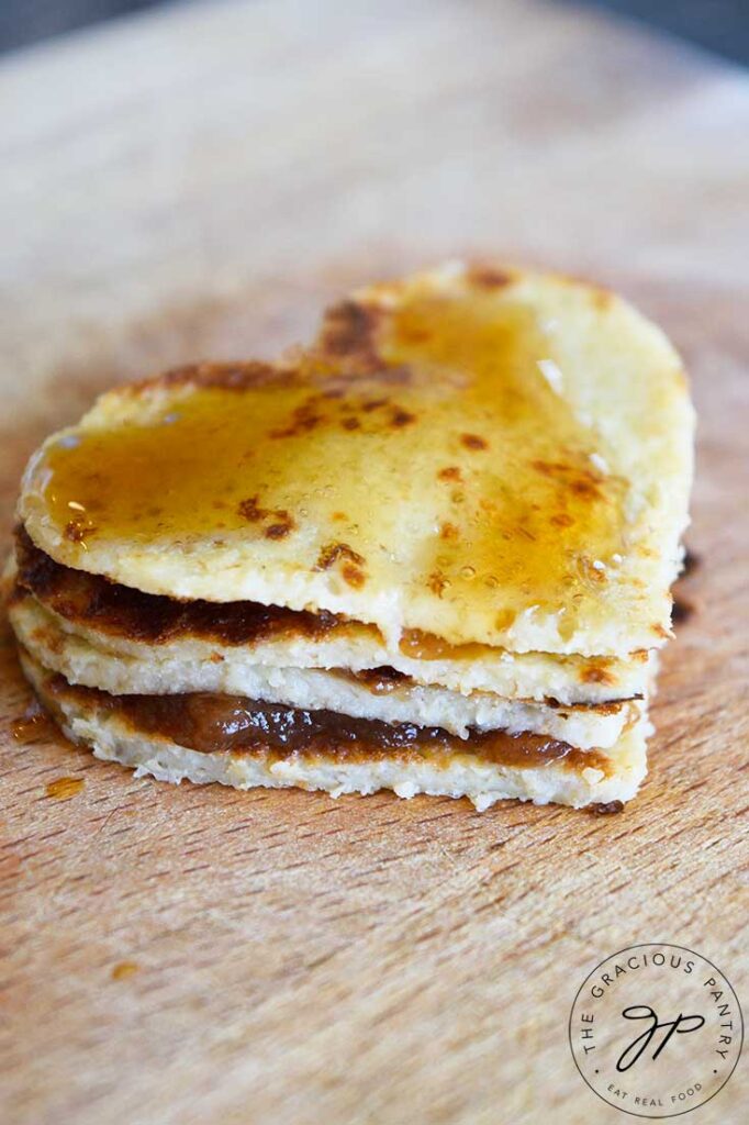 A Pancake Sandwich cut into a heart shape for Valentine's day, with strawberry jam between the 4 layers of heart-shaped pancakes. Maple syrup has been drizzled over the top.