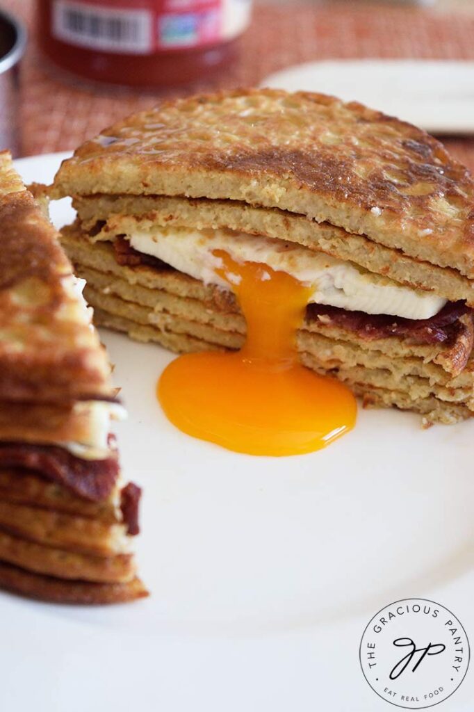 A pancake sandwich sits cut in half. The egg yolk in the center is drizzling down to the center of the plate.