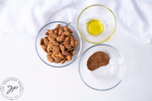 Spicy Roasted Almonds Recipe Ingredients. A small bowl of almonds, a bowl of oil and a bowl of spices sitting next to each other.