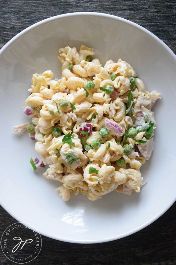 And overhead view looking down into a white pasta bowl filled with this Tuna Pasta Salad. You can see the creamy mayo, green peas, purple onions and a bit of fresh, green garnish sprinkled over the top.