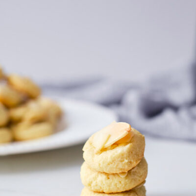 A stack of three Almond Cookies sitting in front of plate filled with more cookies.