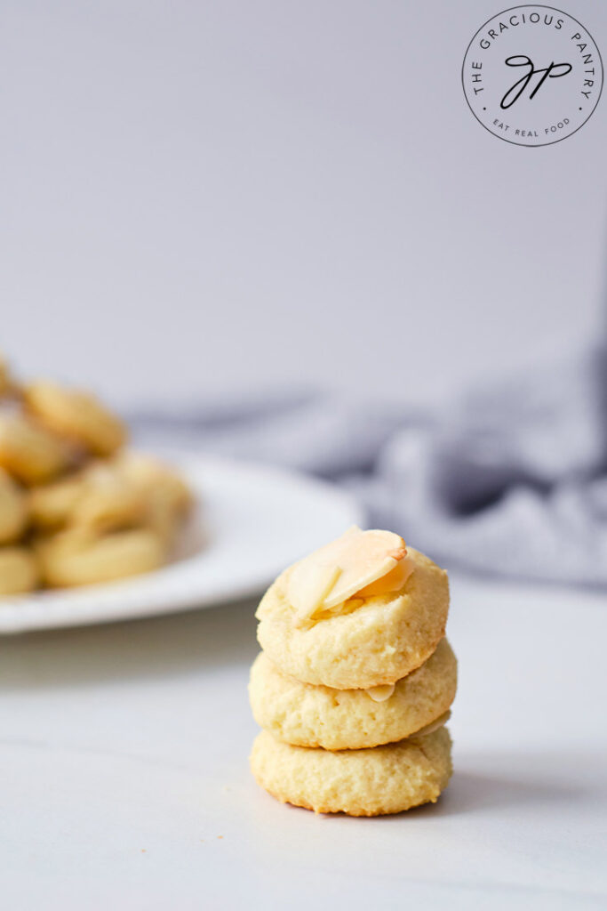 A stack of three Almond Cookies sitting in front of plate filled with more cookies.