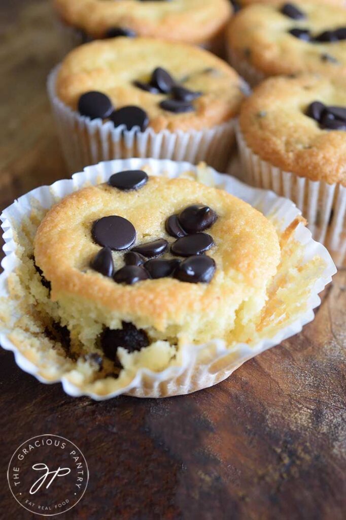 Several almond flour muffins on a wooden surface. The front muffins has the muffin paper peeled away, exposing the bottom half of the muffin.