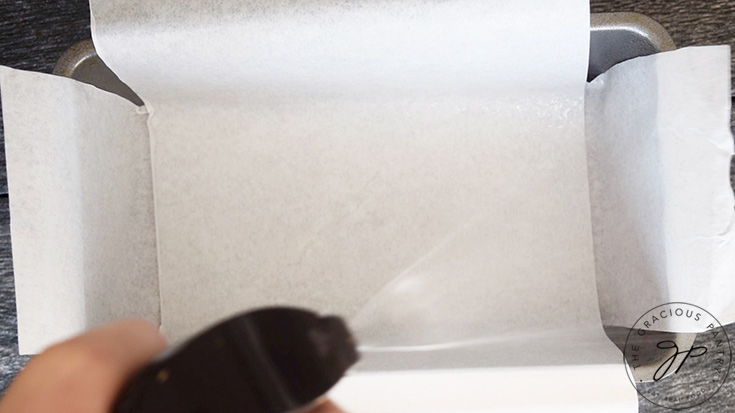 The parchment lined loaf pan being sprayed with oil from an oil sprayer.