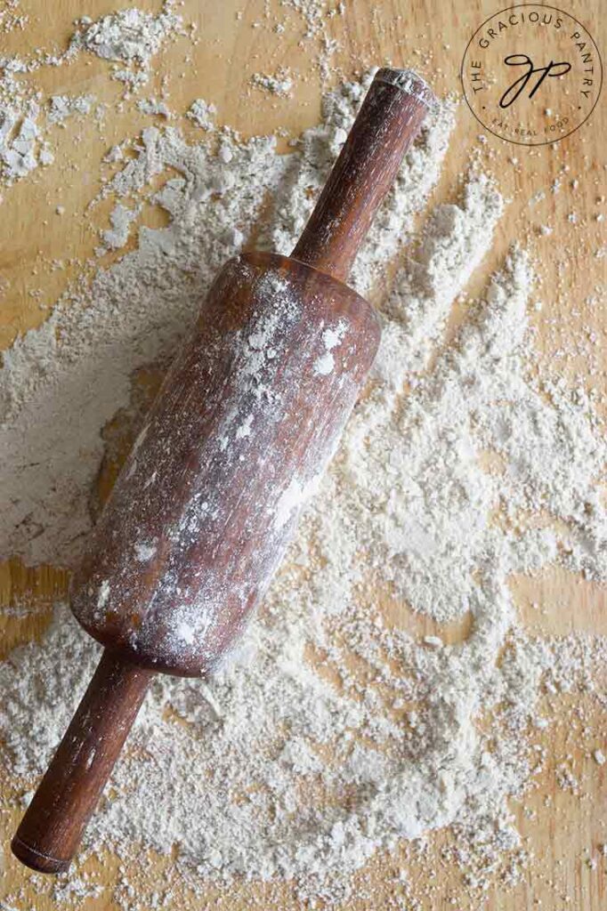 A floured rolling pin sitting on a floured, wooden work surface.