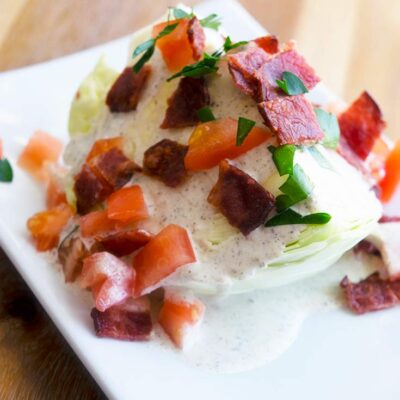 A traditional wedge salad on a white plate with dressing, bacon bits, tomatoes and fresh herbs.