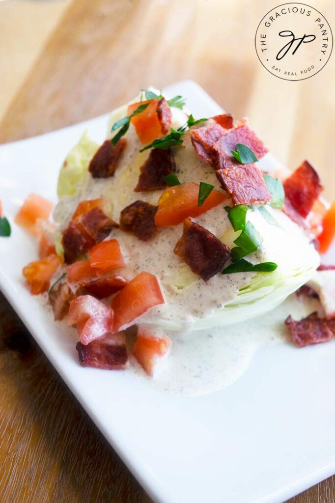 A traditional wedge salad on a white plate with dressing, bacon bits, tomatoes and fresh herbs.