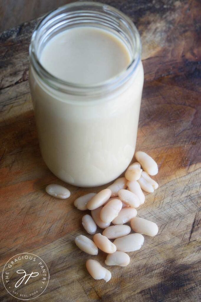 A jar filled with White Bean Alfredo Sauce with a few white beans laying in front of the jar on a wooden surface.