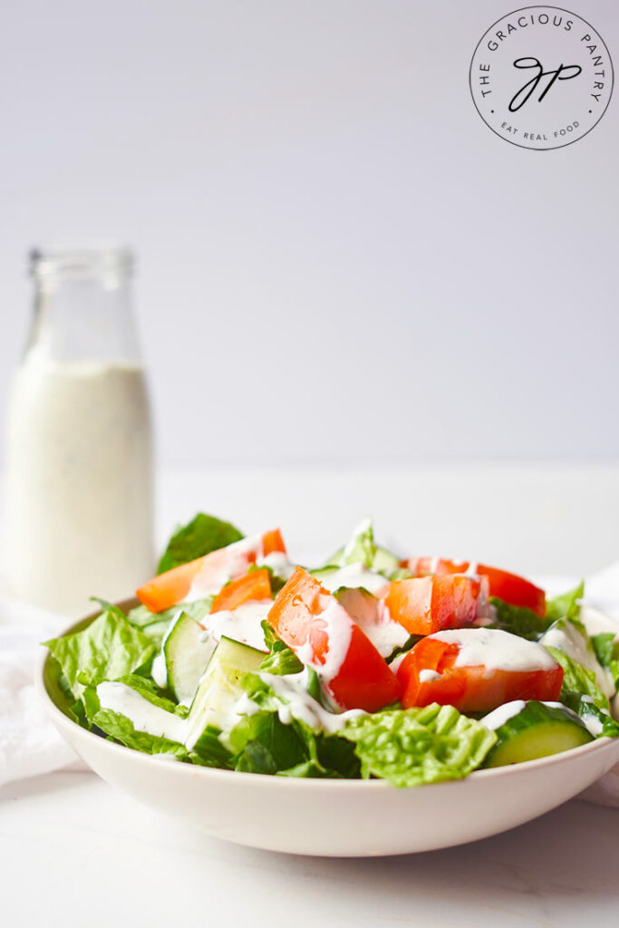 A green salad with cut cucumbers and tomatoes in a white bowl with Buttermilk Ranch Dressing drizzled over the top.