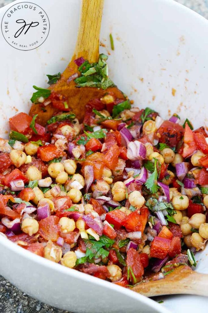 An up close view of this Chickpea Salad in a white mixing bowl with wooden spoons resting in the salad on the side of the bowl.