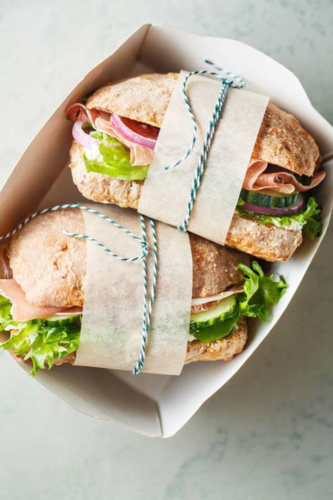 A guide to healthy lunches. Two whole grain sandwiches, wrapped with a parchment strip and ribbon, witting in a sandwich box.
