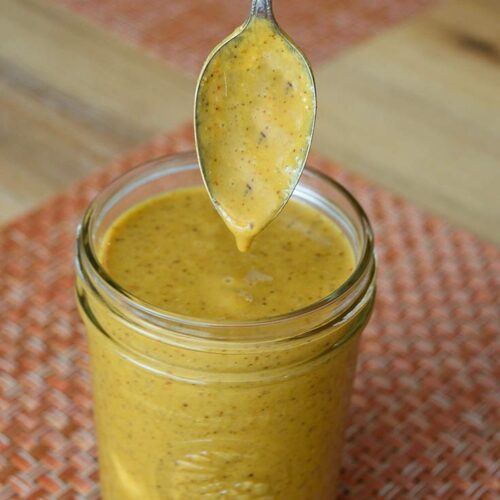 A jar of Carolina Mustard Barbecue Sauce with a spoon just dipped in over the top.