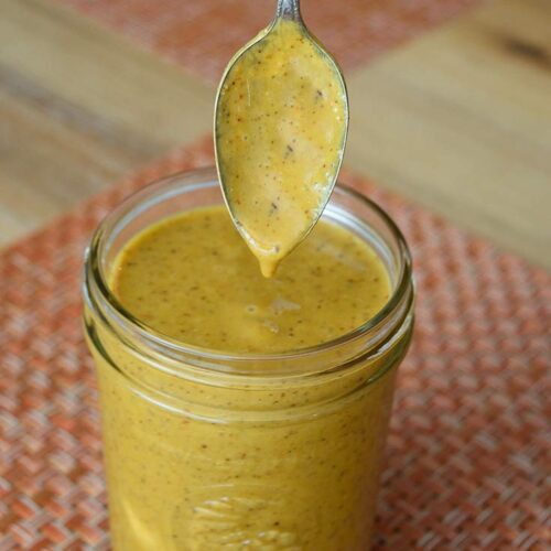 A jar of Carolina Mustard Barbecue Sauce with a spoon just dipped in over the top.