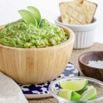 A side view of a wooden bowl filled with guacamole. Lime wedges sit in a small bowl in front.