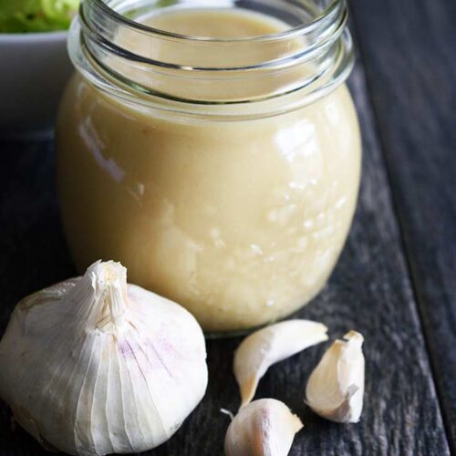 A jar of Roasted Garlic Vinaigrette sits without it's lid on a table. A head of garlic sits in front of it and a bowl of lettuce sits behind it to the left.