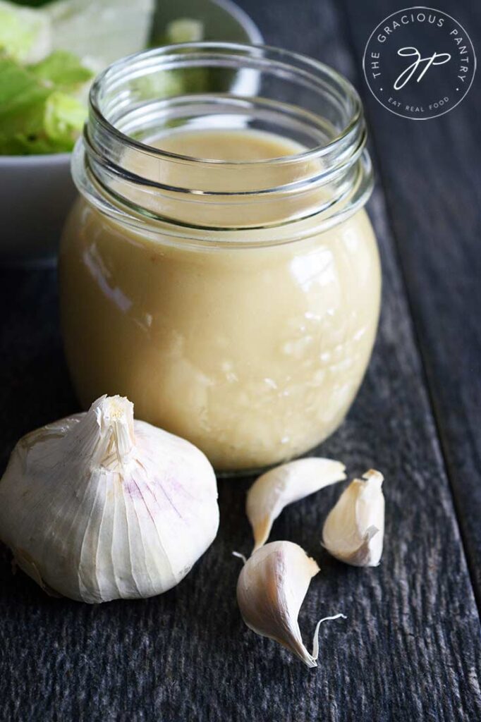 A jar of Roasted Garlic Vinaigrette sits without it's lid on a table. A head of garlic sits in front of it and a bowl of lettuce sits behind it to the left.