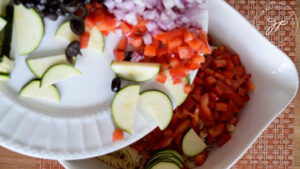 The cut vegetables being added to the cooked pasta in a large, white mixing bowl.