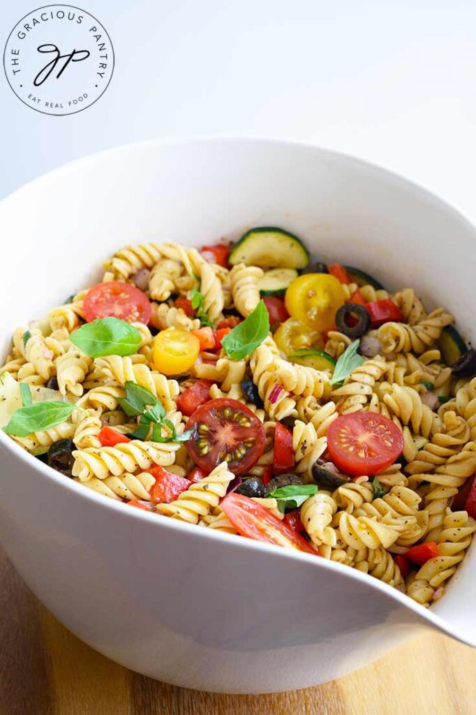 Pasta mixed with tomatoes, zucchini, black olives and fresh basil, fill a white mixing bowl.