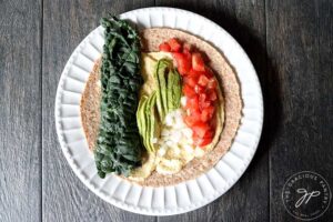 A tortilla with hummus and kale, sliced avocado, chopped onions and tomatoes covering the layer of hummus underneath.