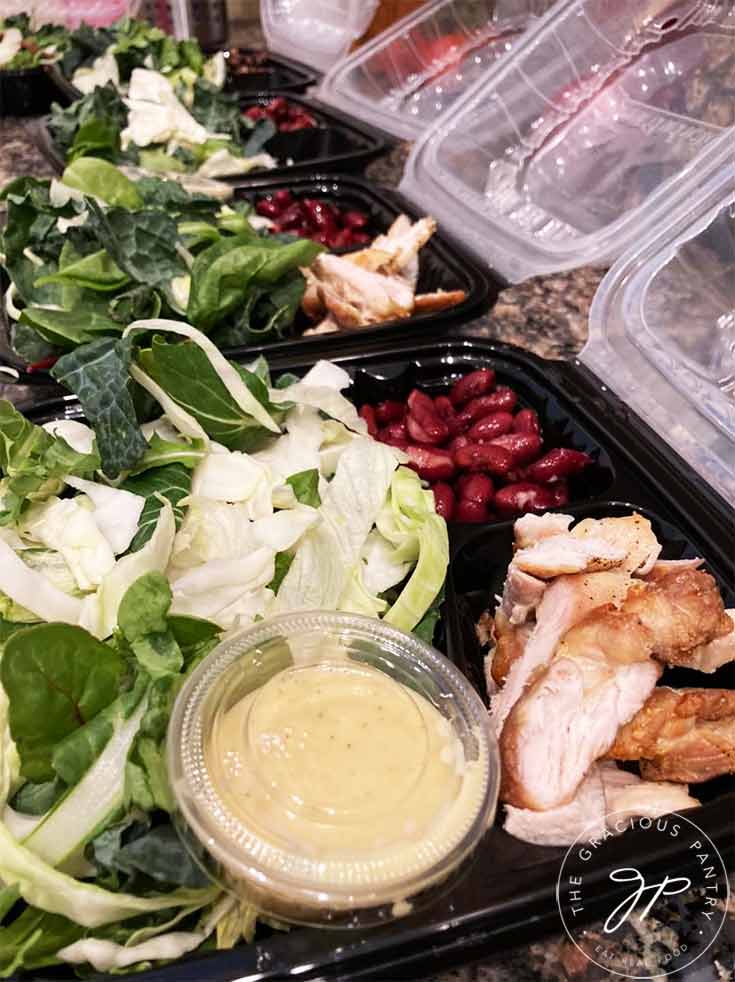 A side view looking down into 2 to-go containers filled with a lettuce mix, kidney beans, sliced chicken and a small container of salad dressing.