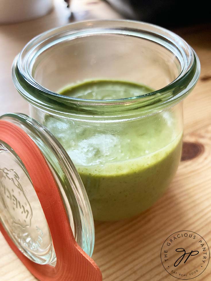 A small canning jar filled half way with this Lemon Basil Dressing Recipe sits on a wooden surface. The jar lid sits propped up against the side of the jar.