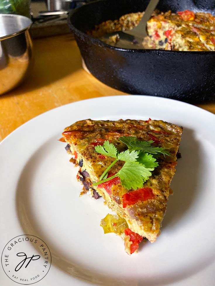 A table view of a single slice of Southwestern Crustless Quiche on a white plate with the cast iron skillet sitting behind it. Other bowls sit in the background.