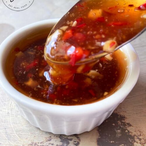 A small, white condiment dish sits filled with this Thai Sweet Chili Sauce. A spoon lifts some of it up for the camera.