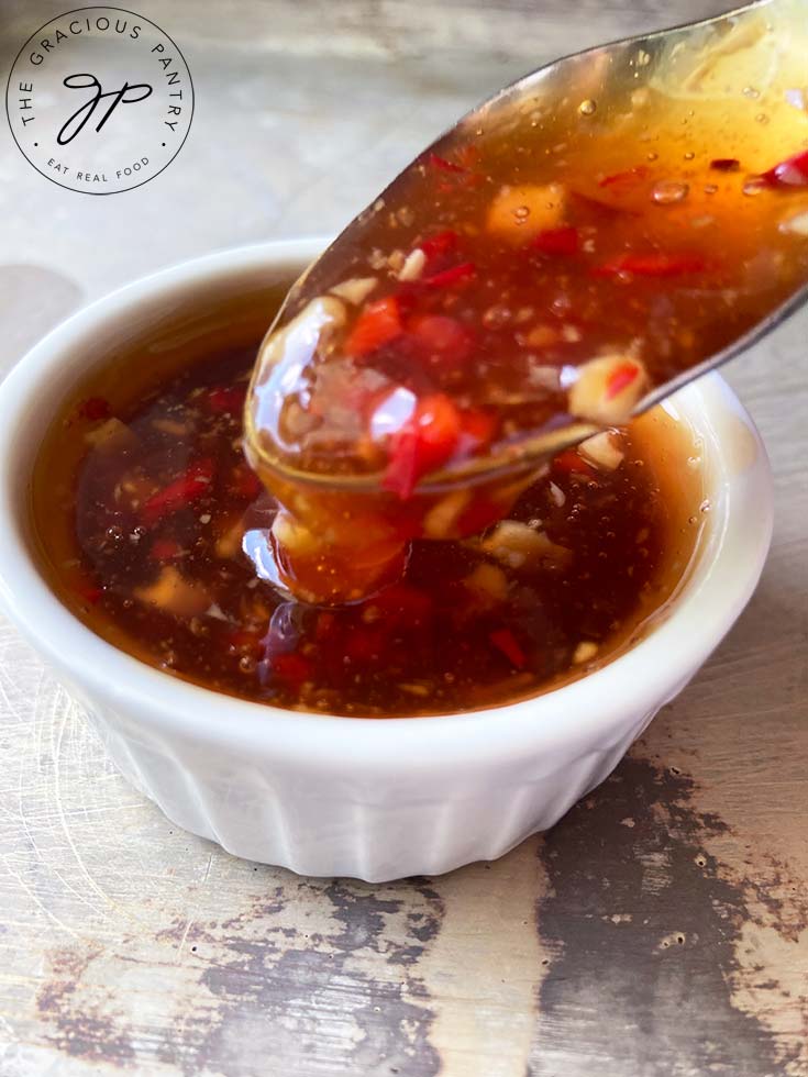 A small, white condiment dish sits filled with this Thai Sweet Chili Sauce. A spoon lifts some of it up for the camera.