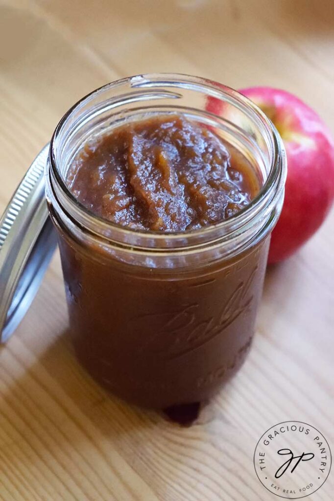 A canning jar filled with this Amish Apple Butter Recipe sits on a wooden surface. The can lid rests on the side of the jar and an apple sits to the back right of the jar.