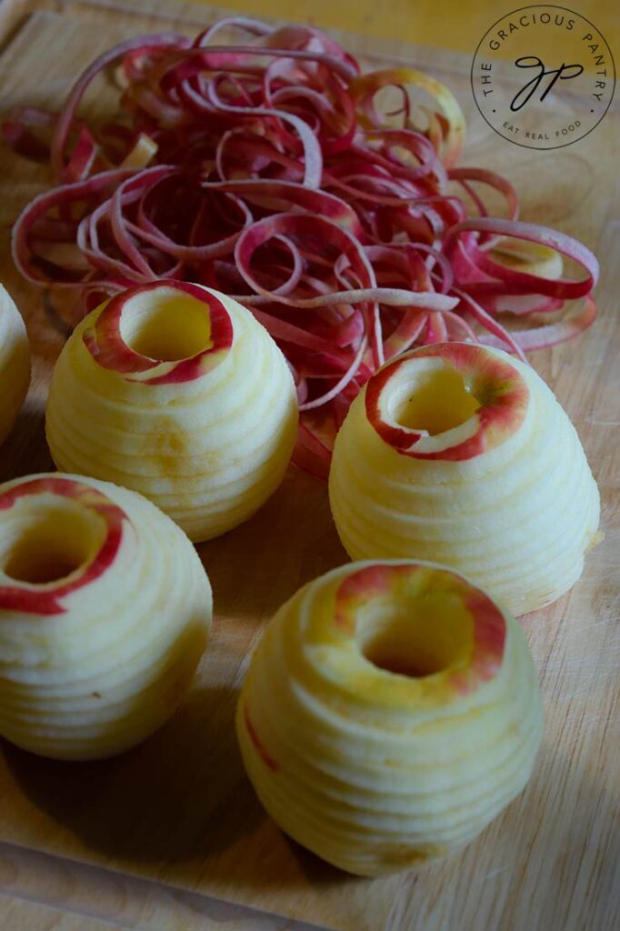 Cored and peeled apples sit on a cutting board waiting to be chopped. A string of apple peel sits behind them.